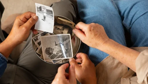 Duas pessoas segurando e olhando fotos antigas em preto e branco guardadas em uma caixa de metal, representando o resgate de memórias e o luto.